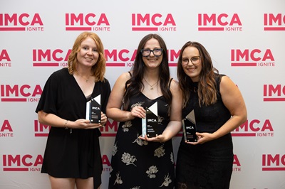 2024_UFG_IMCA_Awards Three women smiling with IMCA backdrop, holding awards.